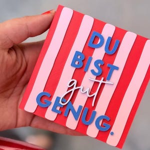 Peut inclure: Une carte carrée avec des rayures verticales rouges et rose clair. La carte présente le texte bleu en relief "DU BIST" en haut, l'écriture cursive blanche "gut" au centre et le texte bleu en relief "GENUG." en bas.