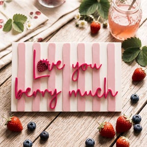 Könnte beinhalten: Ein rechteckiges weißes Schild mit vertikalen hellrosa Streifen und dem erhabenen dunkelrosa Schriftzug "Love you berry much", bei dem ein kleines Erdbeersymbol den Buchstaben "o" in "Love" ersetzt.