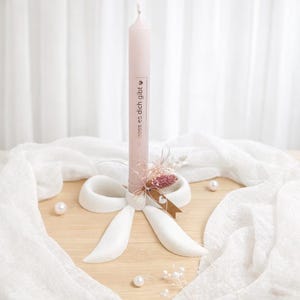 Peut inclure: Une bougie rose clair avec le texte "dass es dich gibt" est présentée sur un support blanc en forme de nœud. La bougie est décorée de fleurs séchées et d'un ruban marron. Le fond comprend du tissu blanc et des perles.