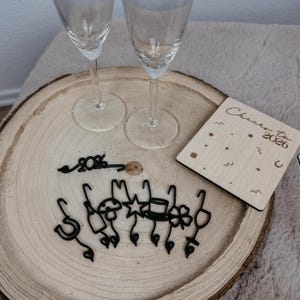 May include: Two champagne flutes sit on a wooden serving tray with a coaster that reads "Cheers to 2026." Black drink charms in various shapes, including a horseshoe, mushroom, and star, are also on the tray.