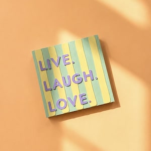 Könnte beinhalten: Ein quadratisches Wandschild mit vertikalen Streifen in abwechselnden Tönen von Hellgrün und Blassgelb. Das Schild zeigt den Text "LIVE. LAUGH. LOVE." in erhabenen, violetten, serifenlosen Großbuchstaben, die mittig auf dem gestreiften Hintergrund angeordnet sind.