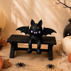 May include: A black bat figurine with large ears, purple cheek spots, and white fangs, sitting on a small black wooden bench. The bat has outstretched wings and dangling legs. The figurine is a decorative item featuring a smooth matte finish.