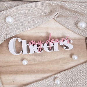 Op de afbeelding: Een houten bord met de tekst "Cheers my dears" in wit en roze. Het bord ligt op een licht houten oppervlak, omgeven door een beige stof en verschillende witte parels.