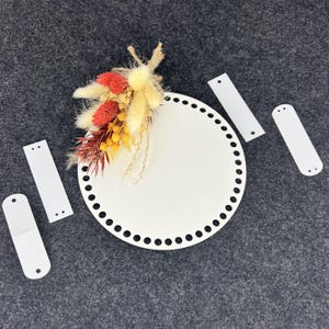 Könnte beinhalten: Eine weiße, runde Holzbasis mit einer Reihe kleiner Löcher am Rand und einem kleinen Strauß getrockneter Blumen in Rot-, Gelb- und Beigetönen. Vier weiße rechteckige Teile mit Löchern sind um die Basis verstreut.