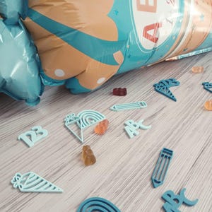 Może przedstawiać: Widok z góry na jasną drewnianą powierzchnię usianą jasnoniebieskimi wycięciami ołówków, tęcz i rożków do lodów. Są też żelki i duży, nadmuchany balon z literami "ABC". Balon jest niebieski i beżowy.