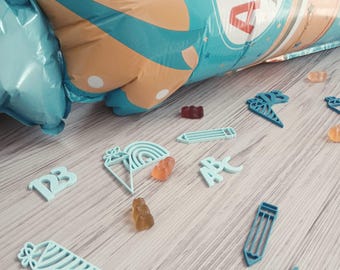 Könnte beinhalten: Eine Draufsicht auf eine helle Holzoberfläche, auf der hellblaue Aussparungen von Bleistiften, Regenbogen und Eiswaffeln verstreut sind. Es gibt auch Gummibärchen und einen großen, aufgeblasenen Ballon mit den Buchstaben "ABC". Der Ballon ist blau und beige.