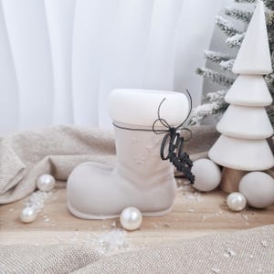 May include: A light gray ceramic Christmas boot with star cutouts and a white cuff. A black ornament with the word "Oh" hangs from the boot. White Christmas tree and pearl ornaments complete the holiday decor.