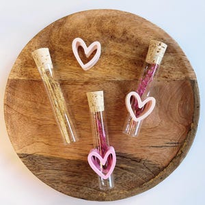 May include: A wooden tray holds three glass vials with cork stoppers, filled with dried flowers and decorative elements. Two vials have pink heart-shaped accents. One vial contains gold-colored strands. A single pink heart is also on the tray.