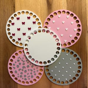 May include: Five round coasters in various colors and patterns. One white coaster with burgundy bows, a pink coaster with white hearts, a pink coaster with gray leopard spots, a white coaster, and a gray coaster with white dots.