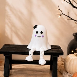 Puede incluir: Una figura de fantasma blanca con un cuerpo liso y redondeado, ojos negros, una pequeña sonrisa y mejillas circulares moradas. El fantasma tiene un pequeño lazo negro en la cabeza y dos piernas blancas articuladas que cuelgan de un banco negro.