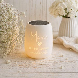 May include: A white solar-powered candle holder with the word "Mama" and a heart symbol, along with the phrase "DU BIST EINFACH WUNDERVOLL" etched on the front. The candle holder has a black top and is surrounded by white flowers.