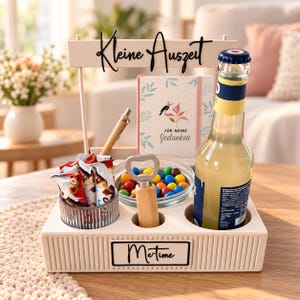 Puede incluir: Una bandeja organizadora rectangular de color beige con un letrero que dice Kleine Auszeit y una etiqueta que dice Me-time. Contiene una botella de vidrio, un cuenco de cristal con caramelos de colores, un abridor, un bolígrafo y un cuaderno.