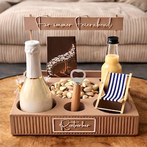 Può includere: Un vassoio da portata beige con scomparti, contenente una bottiglia di champagne, una bottiglia di succo, pistacchi, un apribottiglie, una sedia a sdraio in miniatura e una barretta di cioccolato. Il vassoio riporta la scritta "Far immer Feierabend" e "Rentnerbar".