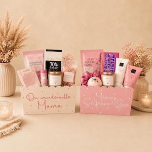 May include: Two rectangular gift boxes containing self-care items. The left box is white with the text 'Du wundervolle Mama' and contains tea, chocolate, and a candle. The right box is pink with the text 'Mamas Selfcare-Box' and contains eye pads, a bath bomb, and skincare tubes.