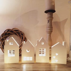 May include: Four white, house-shaped candle holders with cut-out windows and heart designs. The candle holders are illuminated from within, casting a warm glow. A decorative wreath and a candle holder are in the background.
