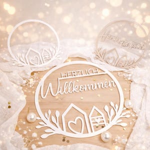Könnte beinhalten: Weiße lasergeschnittene Holzschilder mit den Worten "HERZLICH Willkommen" und "Hereinspaziert" werden angezeigt. Jedes Schild zeigt ein Hausdesign mit Herz- und Blättermotiven. Die Schilder befinden sich auf einer Holzoberfläche mit Perlen und weißem Stoff.