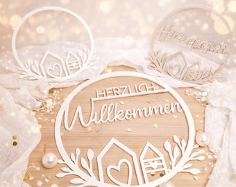 May include: White laser-cut wooden signs with the words "HERZLICH Willkommen" and "Hereinspaziert" are displayed. Each sign features a house design with a heart and leaf accents. The signs are on a wooden surface with pearls and white fabric.