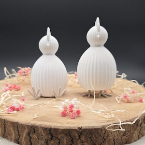 May include: Two white chick ornaments with ribbed bodies and small beaks. They are displayed on a wooden slice, decorated with dried flowers and straw. The figurines have small feet and a vertical crest. The background is dark.
