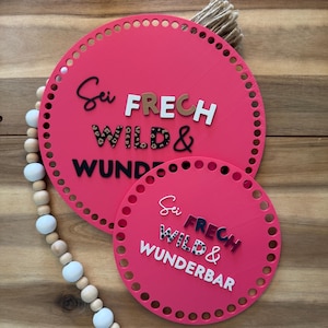 May include: Two pink circular decorative signs with the text "Sei FRECH WILD & WUNDERBAR". The larger sign has a diameter of approximately 10 inches, and the smaller sign is about 6 inches. The signs have a series of holes around the edges.