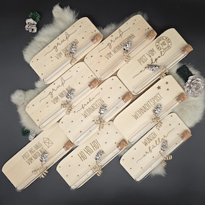 Könnte beinhalten: Eine Auswahl an Holzgeschenkboxen mit verschiedenen Weihnachtsgrüßen und Sternmustern. Jede Box enthält ein Glasröhrchen mit Korken, eine dekorative Tannenzapfen und ein Band. Die Texte auf den Boxen enthalten Sätze wie "Frohe Weihnachten" und "Frohe Feiertage".