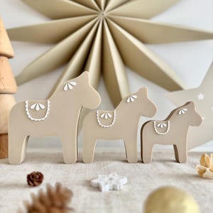 Pode incluir: Três cavalos de brinquedo de madeira em tamanhos variados. Os cavalos são pintados em bege neutro com detalhes brancos na sela. O fundo inclui uma grande estrela e outros elementos decorativos.