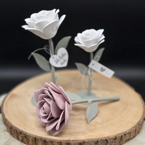 Könnte beinhalten: Drei dekorative Metallrosen mit weißen und malvenfarbenen Blütenblättern, grünen Blättern und Stielen. Zwei Rosen stehen aufrecht, während eine auf einer Holzoberfläche liegt. Jede Rose hat ein kleines Etikett. Der Hintergrund ist dunkel.