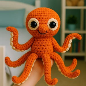 May include: A handmade orange crochet octopus with large black and white eyes and a smiling face. The octopus has eight orange tentacles with cream-colored edges. The toy is held in a person's hand.