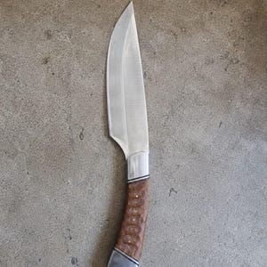 May include: A fixed-blade knife featuring a sharp, pointed steel blade and a curved, textured brown wooden handle. The handle is accented with silver-colored metal bolsters at both ends, and the pommel includes a small circular hole for a lanyard.