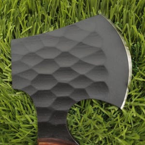 May include: A black axe with a textured head and a sharp, silver blade. The handle is a reddish-brown colour. The axe is resting on a bed of green artificial grass.