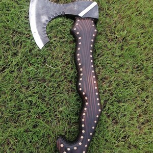 May include: A handcrafted axe with a dark wooden handle decorated with white dot accents. The axe head is metal with a curved blade and a serrated edge. The axe is shown on green grass.