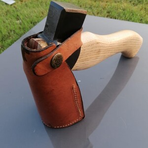 Pu&ograve; includere: Un'ascia artigianale con manico in legno e lama in metallo scuro. La testa dell'ascia &egrave; protetta da un fodero in pelle marrone fissato con un bottone decorativo. Il manico &egrave; in legno chiaro.