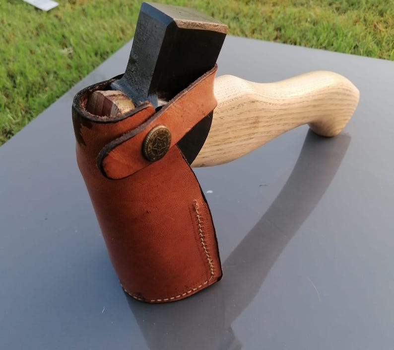 Pu&ograve; includere: Un'ascia artigianale con manico in legno e lama in metallo scuro. La testa dell'ascia &egrave; protetta da un fodero in pelle marrone fissato con un bottone decorativo. Il manico &egrave; in legno chiaro.