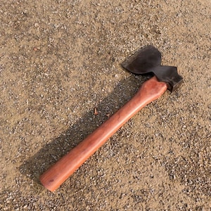 May include: A vintage axe with a dark metal head and a polished wooden handle. The axe head is a dark gray color, and the handle is a warm brown. The axe is lying on a textured, light brown surface.