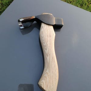 May include: A black axe head with a polished blade and a light-colored wooden handle. The axe is designed for chopping and splitting wood. The handle is curved for a comfortable grip.