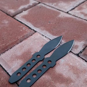May include: Two black throwing knives with pointed blades and a series of circular cutouts in the handles. The knives are lying on a red brick surface, showcasing their sleek, minimalist design. The blades are sharp and ready for use.