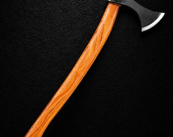 Può includere: Un'accetta con un lungo manico curvo in legno marrone chiaro con venature visibili e una testa in metallo nero opaco con un bordo argentato lucido e affilato. L'attrezzo è posizionato in diagonale su uno sfondo scuro e strutturato.