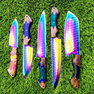 Op de afbeelding: Vijf keukenmessen met iriserende bladen en houten handvatten. De bladen tonen een regenboog aan kleuren, terwijl de handvatten verschillende patronen hebben. De messen zijn gerangschikt op groen gras.