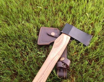 Può includere: Un'accetta con una testa in metallo nero lucido e un manico in legno chiaro con venature visibili. L'accetta è accompagnata da un fodero in pelle marrone scuro con cuciture a vista e una chiusura con bottone a pressione metallico.