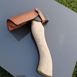 Pu&ograve; includere: Ascia con manico in legno chiaro e lama nera, protetta da un fodero in pelle marrone. L'impugnatura presenta un design curvo per l'impugnatura. Il fodero &egrave; fissato con un cinturino in pelle e un bottone metallico.