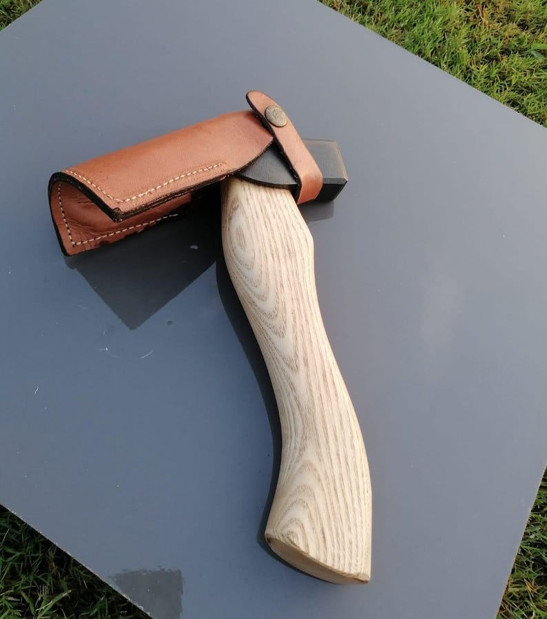Pu&ograve; includere: Ascia con manico in legno chiaro e lama nera, protetta da un fodero in pelle marrone. L'impugnatura presenta un design curvo per l'impugnatura. Il fodero &egrave; fissato con un cinturino in pelle e un bottone metallico.