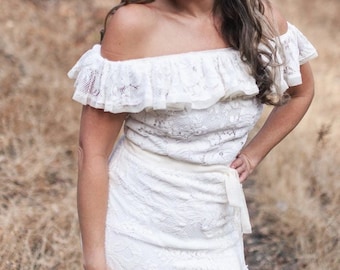 May include: A woman wearing a white off-the-shoulder lace dress with a ruffled neckline and a white flower in her hair. She is standing in a wooded area with dry grass and trees in the background.