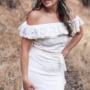 Puede incluir: Una mujer con un vestido blanco de encaje sin hombros con un escote con volantes y una flor blanca en el pelo. Est&aacute; de pie en una zona boscosa con hierba seca y &aacute;rboles en el fondo.