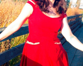 May include: A woman wearing a red dress with white lace trim and a fitted bodice. The dress has a full skirt and is flowing in the wind. She is standing on a wooden walkway.