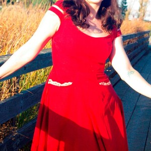 Puede incluir: Una mujer con un vestido rojo con ribetes de encaje blanco y un corpi&ntilde;o ajustado. El vestido tiene una falda completa y ondea con el viento. Est&aacute; de pie en una pasarela de madera.