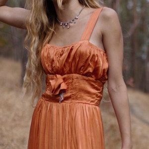 May include: A woman wearing a long, orange, sleeveless dress with a cinched waist and a bow detail. She is standing on a path in a forest.
