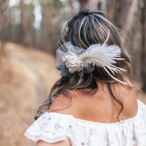 Puede incluir: Una mujer con el pelo largo y oscuro que lleva un vestido blanco de encaje de hombros descubiertos con un accesorio para el pelo de flor blanca y pluma.