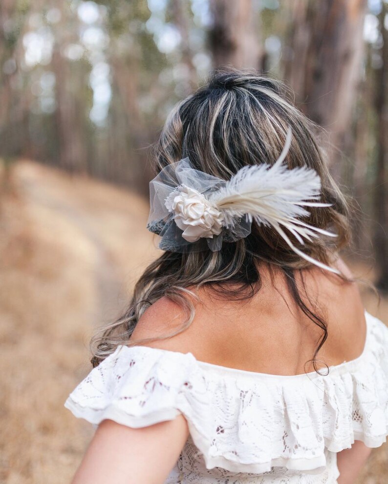 Puede incluir: Una mujer con el pelo largo y oscuro que lleva un vestido blanco de encaje de hombros descubiertos con un accesorio para el pelo de flor blanca y pluma.