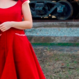 Puede incluir: Una mujer con un vestido rojo con ribete de encaje blanco. El vestido tiene un corpi&ntilde;o ajustado y una falda completa. La mujer est&aacute; de pie frente a una v&iacute;a de tren.