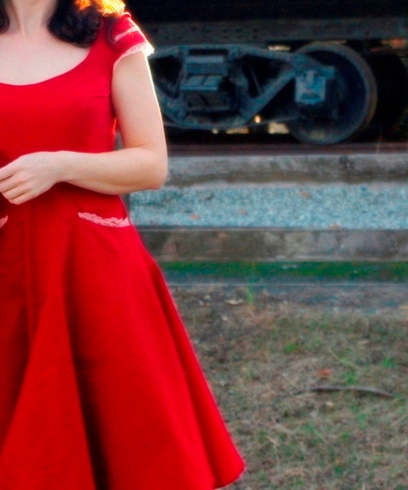 Puede incluir: Una mujer con un vestido rojo con ribete de encaje blanco. El vestido tiene un corpi&ntilde;o ajustado y una falda completa. La mujer est&aacute; de pie frente a una v&iacute;a de tren.