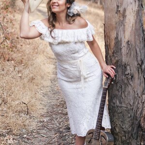 Puede incluir: Una mujer con un vestido blanco de encaje se encuentra en un &aacute;rea boscosa, sosteniendo un sombrero en una mano y una guitarra en la otra. La guitarra es una guitarra resonadora con un agujero de sonido redondo y un cuerpo de metal.